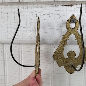 May include: Two ornate brass-colored metal wall hooks. One hook is fully visible, the other is partially visible. The hooks have a decorative design and a black curved hook. The hooks are mounted on a white, distressed wooden surface.