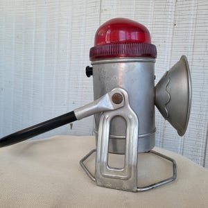 May include: Vintage bicycle lamp with a red lens and a silver metal body. The lamp features a black handle and a decorative front reflector. The base is a metal frame. A classic piece of cycling equipment.