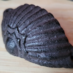 May include: A black cast iron drawer pull with a scalloped design. The pull has a hole on the side for mounting.