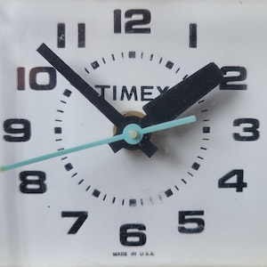 May include: Close-up of a white Timex analog clock face. Black hands point to the time, with black numerals marking the hours. The second hand is a light blue color. The words "TIMEX" are printed in black. The clock is marked "MADE IN U.S.A."