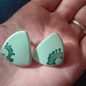 May include: A pair of light blue guitar picks with a green and white design. The picks are shaped like a triangle with rounded edges.