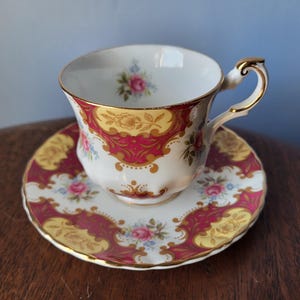 May include: A vintage teacup and saucer set. The cup and saucer are white with a gold trim and a decorative pattern in red and yellow. The set features floral accents in pink and blue.