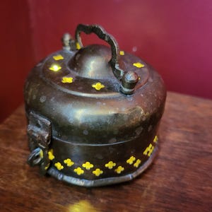 Peut inclure: Une petite boîte à bijoux ronde en laiton avec un couvercle à charnière et une poignée décorative. La boîte est décorée d'un motif d'étoiles jaunes.