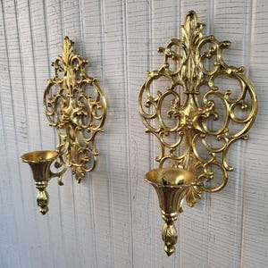 May include: A pair of gold-colored metal wall sconces featuring intricate scrollwork and filigree patterns. Each sconce has a central vertical design with a flared, cup-shaped candle holder at the base, supported by a decorative, tapered stem.