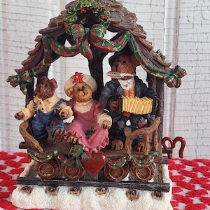 Può includere: Una statuetta decorativa con tre orsacchiotti su un vagone ferroviario sotto un tetto decorato con nastri a quadri verdi e rossi. Un orso indossa una salopette blu, un altro un vestito rosa e il terzo un abito scuro con una fisarmonica gialla.