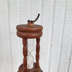 May include: A wooden hourglass with a dark brown finish, featuring four turned pillars and a small, carved wooden mouse figure with a long tail perched on the top circular platform. The hourglass contains white sand within a clear glass bulb.