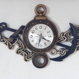 May include: A nautical-themed wall clock featuring a round clock face with a compass design, surrounded by a bronze-colored frame. Two black anchors and chains flank the clock. The clock hands are black.