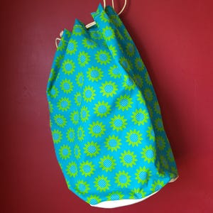 May include: A drawstring bag with a bright turquoise background and a repeating pattern of green sunflowers with white centers. The bag is hanging against a red wall.