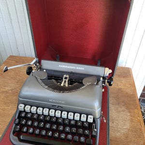 May include: A grey Remington Rand portable typewriter with a QWERTY keyboard featuring black and white keys. The typewriter is housed in a dark grey carrying case with a vibrant red interior lining. The brand name Remington is printed on the front.