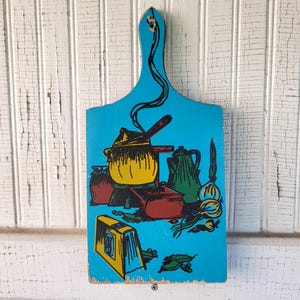 May include: A vintage, blue wooden cutting board with a handle, featuring a colorful kitchen-themed illustration. The artwork includes a yellow pot, red pots, a green teapot, and onions. The board has a hole for hanging.
