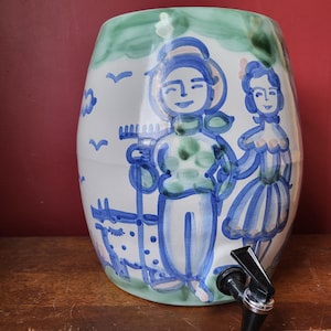 Puede incluir: Un dispensador de agua de cerámica con un diseño pintado a mano. El dispensador blanco presenta ilustraciones azules y verdes de figuras y elementos paisajísticos. Tiene un grifo negro y un borde verde.