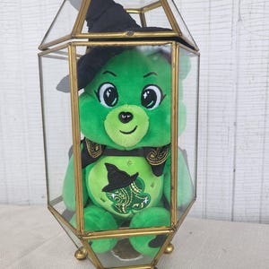 May include: A green plush bear wearing a black witch hat and a decorative scarf, enclosed in a clear glass and brass terrarium. The bear has a witch hat and paisley design on its belly. The terrarium has a geometric shape and sits on small brass feet.