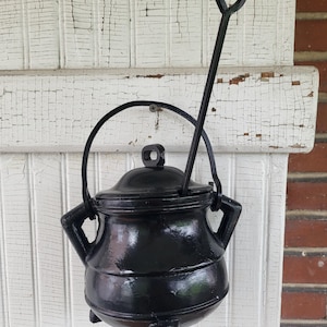 May include: A black cast iron cauldron with a matching lid, a curved handle, and three short legs. A long, thin metal rod with a circular loop at the top extends vertically from the side of the pot, providing a structure for hanging or display.