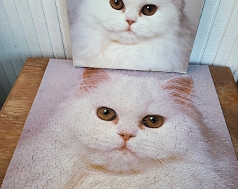 Puede incluir: Un rompecabezas cuadrado que presenta el retrato de un gato blanco esponjoso con grandes ojos color ámbar, exhibido junto a la tapa de su caja original que muestra la misma imagen de alta resolución del felino de pelo largo.