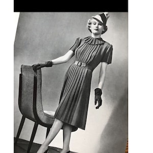 May include: A black and white photo of a woman wearing a pleated dress and a hat. She is standing next to a chair with her hand on the back of the chair.