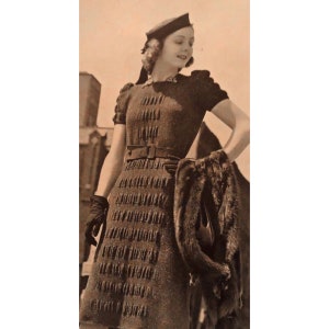 Può includere: Una fotografia in bianco e nero di una donna che indossa un abito di maglia marrone con una cintura, un berretto nero e guanti neri. Tiene una stola di pelliccia nella mano destra.