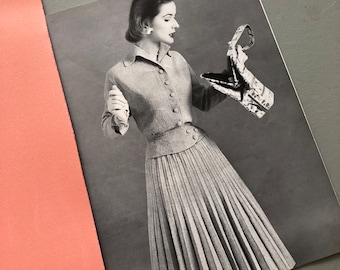 May include: A black and white photo of a woman wearing a pleated skirt and a button-up sweater. She is holding a small, patterned handbag.