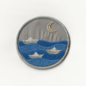 May include: Embroidered patch with a gray background and blue waves. Three white paper boats are floating on the waves. A crescent moon and stars are in the sky.