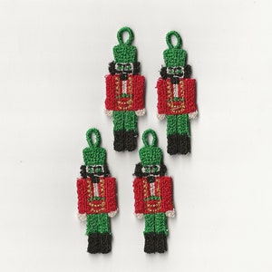 May include: Four green and red felt nutcracker ornaments with gold accents. The ornaments are hanging from a white background.