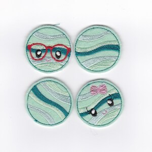 May include: Four round, light blue felt patches with embroidered designs. Two patches have a wavy pattern in teal and silver. One patch has a red pair of glasses and two black eyes. The other patch has a pink bow and two black eyes.