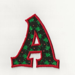 Peut inclure: Écusson brodé en forme de lettre "A" avec un fond noir orné d'illustrations de grenouilles vertes et d'étoiles rouges. L'écusson a une bordure rouge. Ce patch décoratif peut être cousu ou repassé sur du tissu.