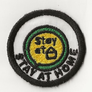 Puede incluir: Un parche bordado en blanco y negro con el texto "Stay at Home" en un círculo. El centro del parche tiene un círculo amarillo con un contorno verde y el texto "Stay at" dentro. Un icono de casa está en el centro del círculo amarillo.
