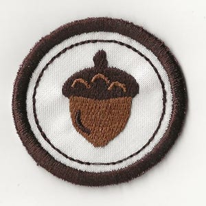 May include: A circular embroidered patch featuring a brown acorn design on a white background. The acorn is detailed with brown stitching, and the patch has a brown border. The patch is likely for clothing or accessories.