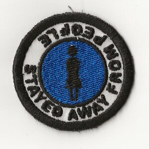 Puede incluir: Un parche bordado en blanco y negro con un círculo azul en el centro. El círculo tiene una silueta negra de una persona de pie en el centro. El texto alrededor del círculo dice "Stay Away From People".