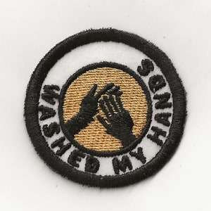 May include: A black and white embroidered patch with a gold background. The patch is round and has the text "Washed My Hands" written around the edge. Two hands are shown in the center of the patch, with one hand washing the other.