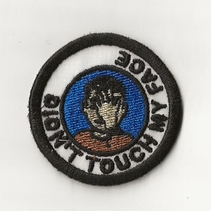 May include: A black and white embroidered patch with a blue circle in the center. The circle has a cartoon image of a person with their hands over their face. The text around the circle reads "Don't Touch My Face".