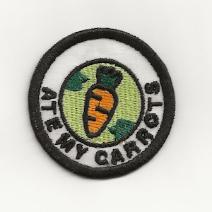May include: A round, black and white embroidered patch with a green and orange carrot in the center. The text "ATE MY CARROTS" is written around the carrot in a black circle.