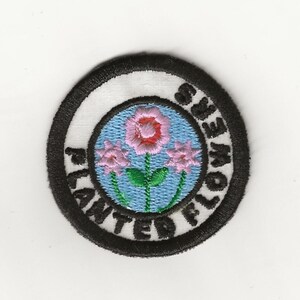 May include: A black and white embroidered patch with a blue circle in the center. The circle has three pink flowers with green stems and leaves. The text "Planted Flowers" is written around the circle.