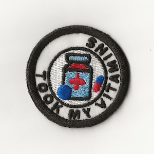 May include: Embroidered patch with a black border and white background. The patch features a blue and red pill bottle with a red cross, a blue sphere, and a blue and red capsule. Text around the edge reads "I TOOK MY VITAMINS".