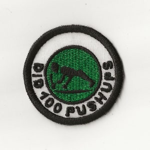 May include: A black and white embroidered patch with a green circle in the center. The circle features a silhouette of a person doing a push-up. The text around the circle reads "Did 100 Pushups".