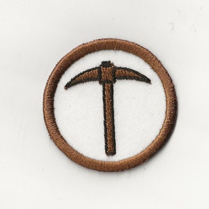 Puede incluir: Un parche bordado marrón con un círculo blanco en el centro. El círculo contiene un contorno negro de un pico.