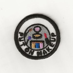 May include: A black and white embroidered patch with a white background and a black circle around the edge. The circle contains a gray makeup palette with colorful makeup items and the text "Put on Makeup" in black.