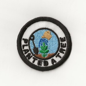 May include: A black and white embroidered patch with a circle design. The center of the circle shows a green plant with a brown pot pouring water. The text "Planted a Tree" is written around the circle.
