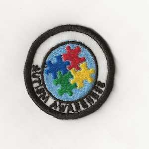 May include: A round, black and white embroidered patch with the text "Autism Awareness" surrounding a blue circle with four puzzle pieces in red, blue, green, and yellow.