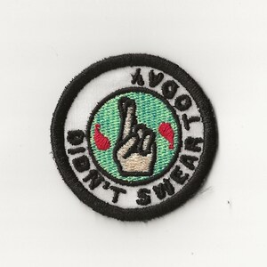 May include: A black and white embroidered patch with a green circle in the center. The circle has a white hand with two fingers crossed and the text "I  DIDN'T SWEAR TO  OBEY" around the outside.