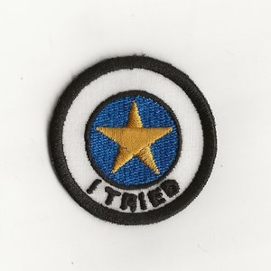 Puede incluir: Un parche redondo bordado en blanco y negro con un círculo azul en el centro. El círculo tiene una estrella dorada en el centro y las palabras "I TRIED" alrededor.