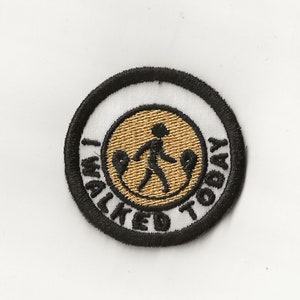 May include: A round, white embroidered patch with a black border. The center of the patch features a gold circle with a black stick figure walking. The text "I WALKED TODAY" is written in black around the gold circle.