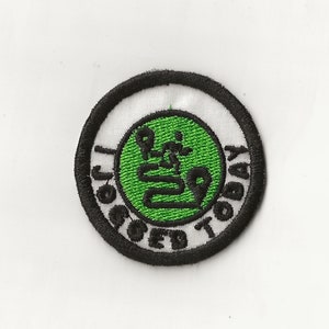 Könnte beinhalten: Ein gestickter Aufnäher in Schwarzweiß mit einem grünen Kreis in der Mitte. Der Kreis enthält ein stilisiertes Bild einer Person, die Yoga macht. Der Text "Get Your Om On Today" ist um den Kreis herum geschrieben.