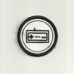 May include: A black and white embroidered patch featuring a retro video game controller design. The controller is outlined in black with a red button and black buttons.