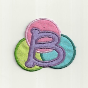 Op de afbeelding: Een paarse viltletter "B" omringd door drie cirkels van roze, groene en blauwe vilt.