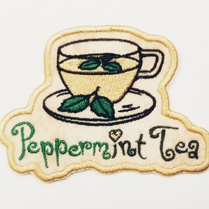 Könnte beinhalten: Gestickter Bügel-Patch mit einer gelben Teetasse und Untertasse mit grünen Blättern und dem Text "Peppermint Tea" in grüner Kursivschrift.