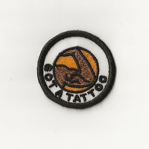 May include: A black and white embroidered patch with a brown and orange circle in the center. The circle has a stylized image of a tattoo needle. The text "Got a Tattoo" is written around the circle.