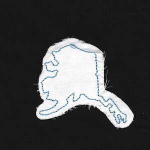 May include: A white fabric patch in the shape of the state of Alaska, outlined in blue thread. The fabric is frayed around the edges.