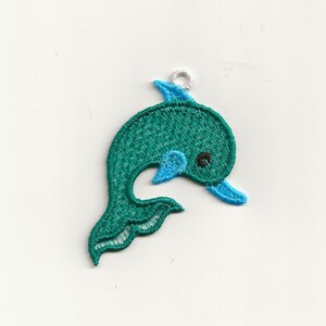 May include: A teal blue embroidered dolphin charm with a white loop for hanging. The dolphin has a blue fin and a black eye.
