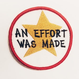 Op de afbeelding: Een ronde, rood omrande patch met een gouden ster en de tekst "AN EFFORT WAS MADE" in zwart.