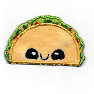 Pode incluir: Um taco de feltro amarelo com um recheio bordado verde e vermelho e um rosto sorridente. O taco tem olhos pretos e uma boca branca.